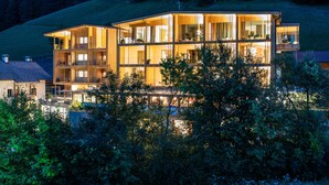 A modern multi-story building with large windows and balconies, surrounded by trees and set on a hillside. - Naturhotel Rainer (Racines)