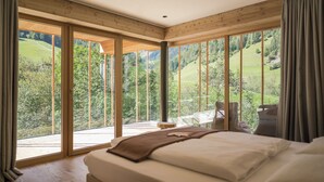 A bedroom with a large bed, a view of a green landscape, and a sliding glass door leading to an outdoor area. - Naturhotel Rainer (Racines)