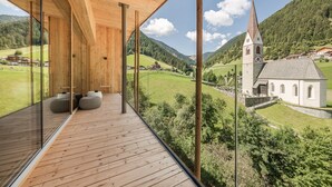 A modern wooden balcony with large windows overlooking a lush valley and a church. - Naturhotel Rainer (Racines)