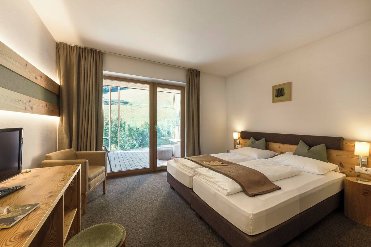A hotel room with a large bed, a desk with a chair, a television, and a view of greenery through the window.