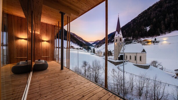 A wooden interior with a large window overlooking a snowy landscape and a church. - Naturhotel Rainer (Racines)