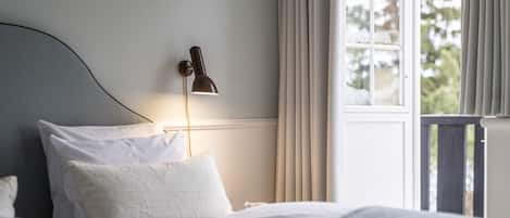 A neatly made bed with white linens, a bedside lamp, and a window with curtains.