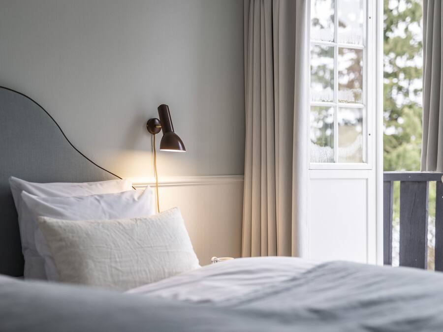 Una cama bien hecha con sábanas blancas, una lámpara de noche y una ventana con cortinas.