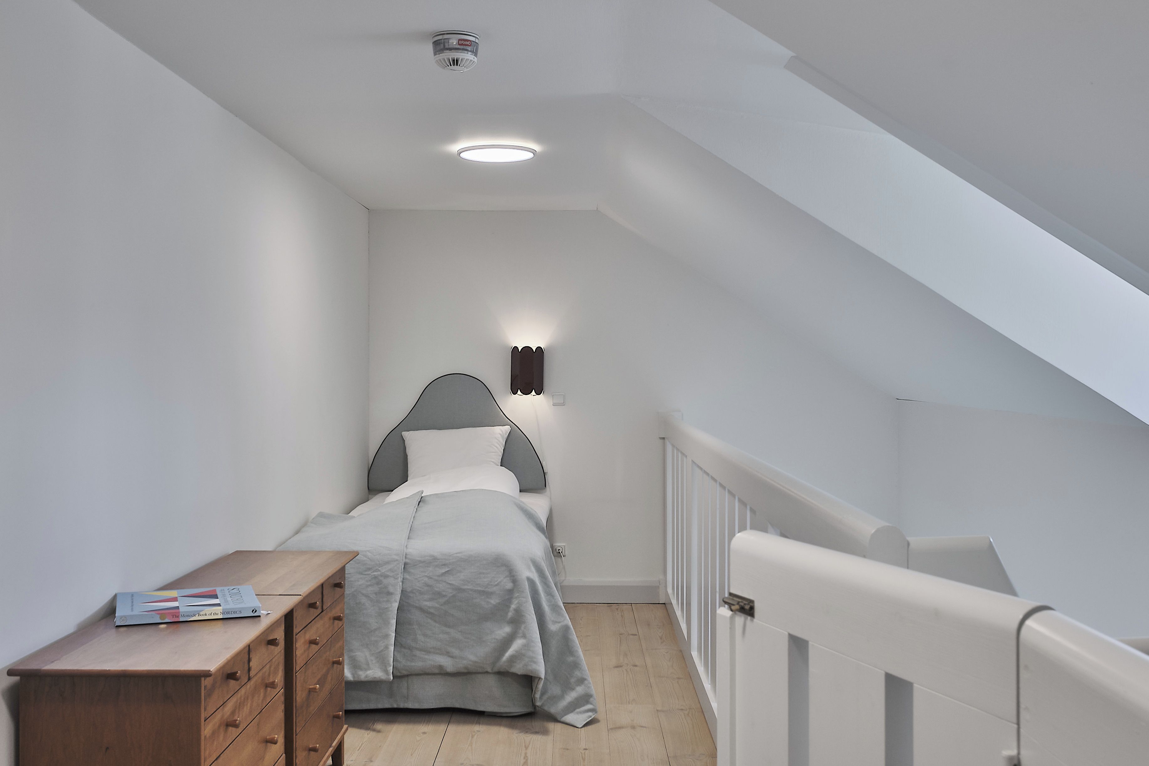 Habitación estrecha y con techo inclinado, con una cama, una mesita de noche y un libro a la izquierda.
