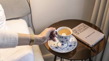 Una mano sostiene una taza de café sobre un platillo, con un libro titulado 'The Little Book of Fashion' sobre una mesa redonda pequeña.