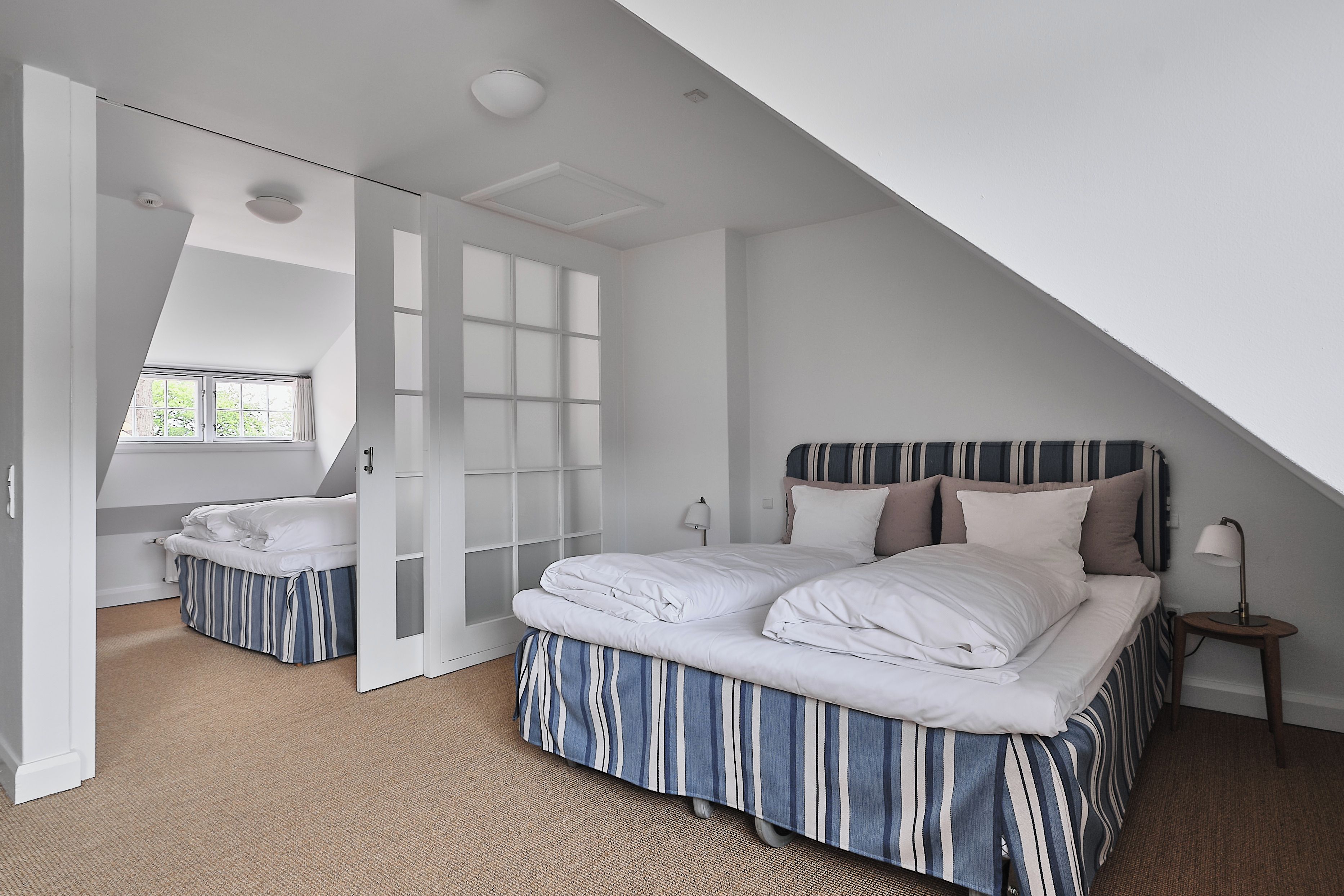 A bedroom with a sloped ceiling, a large window, a bed with striped bedding, a bedside table with a lamp, and a door leading to another room.