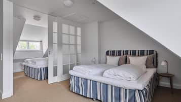 A bedroom with a sloped ceiling, a large window, a bed with striped bedding, a bedside table with a lamp, and a door leading to another room.