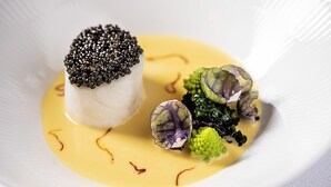 Une assiette blanche sur laquelle repose un poisson blanc, garni de caviar noir, de brocolis verts et de chou-fleur violet.