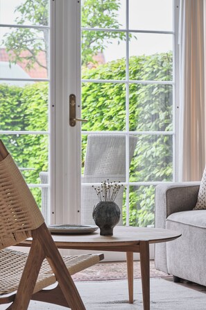 A room with a large window, a wicker chair, a sofa, a table with a vase, and a door with a brass handle. - Molskroen (Ebeltoft)