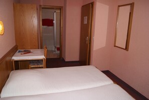 Een kleine hotelkamer met een eenpersoonsbed, een houten garderobe, een tafeltje met een stoel en een deur naar een badkamer.