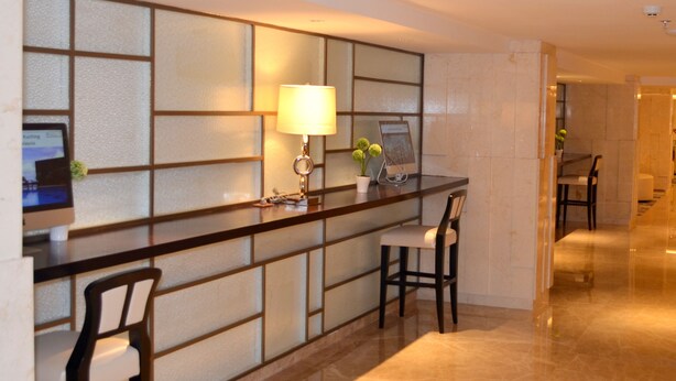 A reception desk with a computer monitor, a lamp, and a potted plant. There are two chairs in front of the desk. The floor is tiled, and the walls have a geometric pattern.
