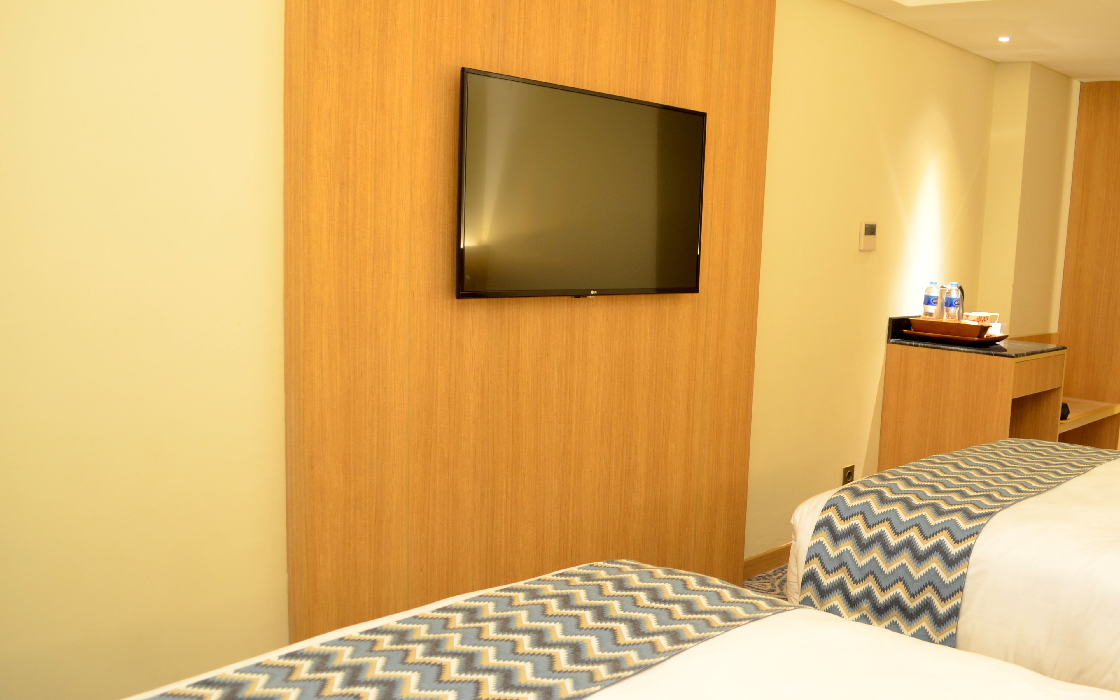 A hotel room with two beds, a flat-screen TV mounted on the wall, a small desk with a water bottle, and a mirror.
