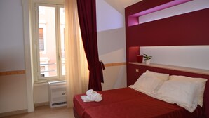 A hotel room with a bed, a window with curtains, a radiator, and a small shelf with a plant. - B&B The Queens (Foggia)