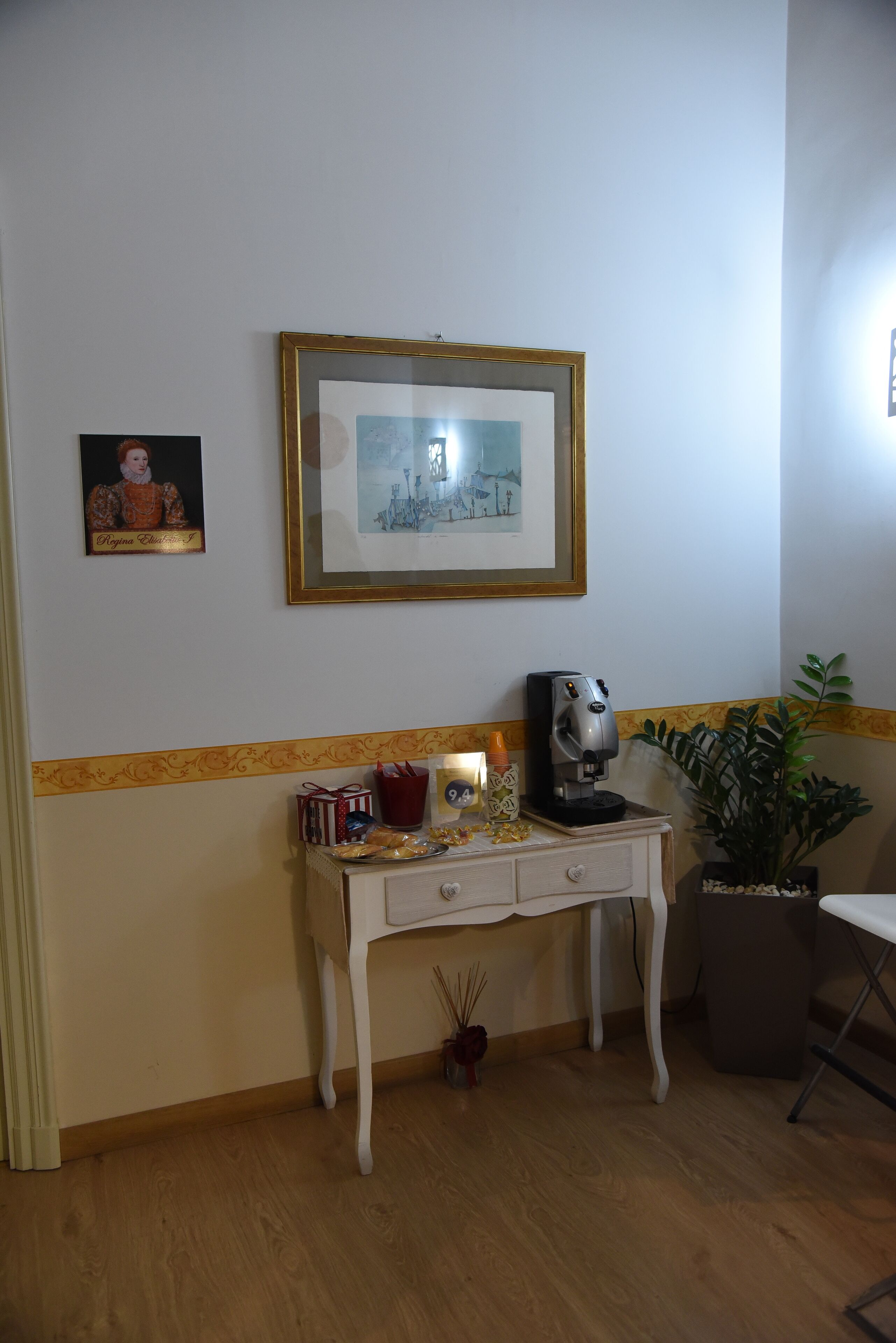 Una habitación con un escritorio blanco, un cuadro enmarcado, una cafetera, una planta en maceta y una pequeña mesa con una vela.