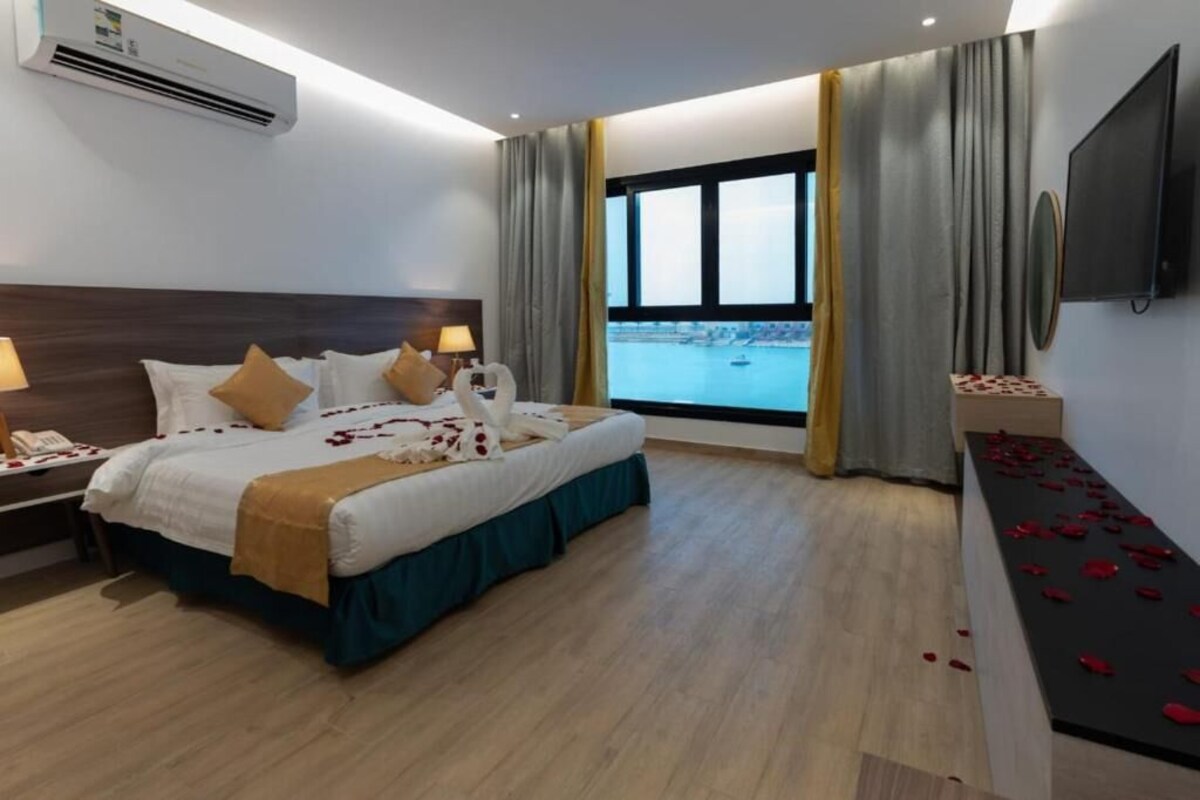 Ein Hotelzimmer mit einem großen Bett, einem Nachttisch, einem Fernseher und einem Fenster mit Blick auf ein Gewässer.