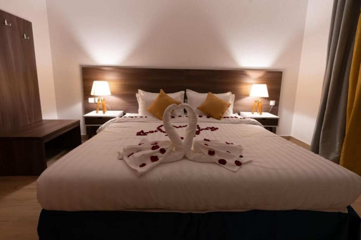 Ein ordentlich bezogenes Bett mit Handtuch und Rosenblättern, die in Herzform angeordnet sind, zwei Nachttischlampen und ein hölzernes Kopfende.