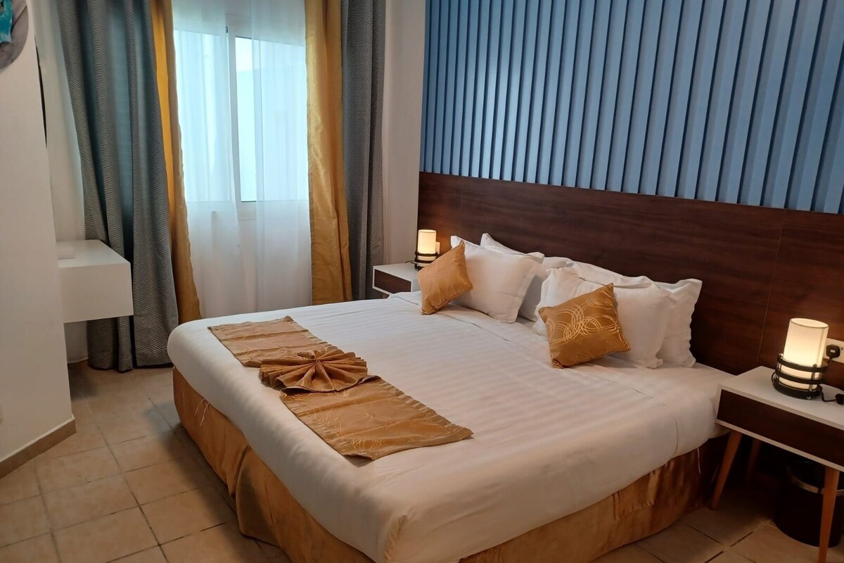 Ein Hotelzimmer mit einem großen Bett, einem Holz-Kopfteil, zwei Nachttischen mit Lampen, einem Schreibtisch und einem Fenster mit Vorhängen.