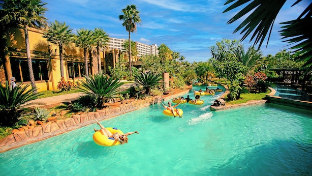 A pool area with multiple people on inflatable tubes, surrounded by palm trees and a building with large windows. -  Hot Beach Raízes (Olímpia)