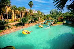 A pool area with multiple people on inflatable tubes, surrounded by palm trees and a building with large windows. -  Hot Beach Raízes (Olímpia)