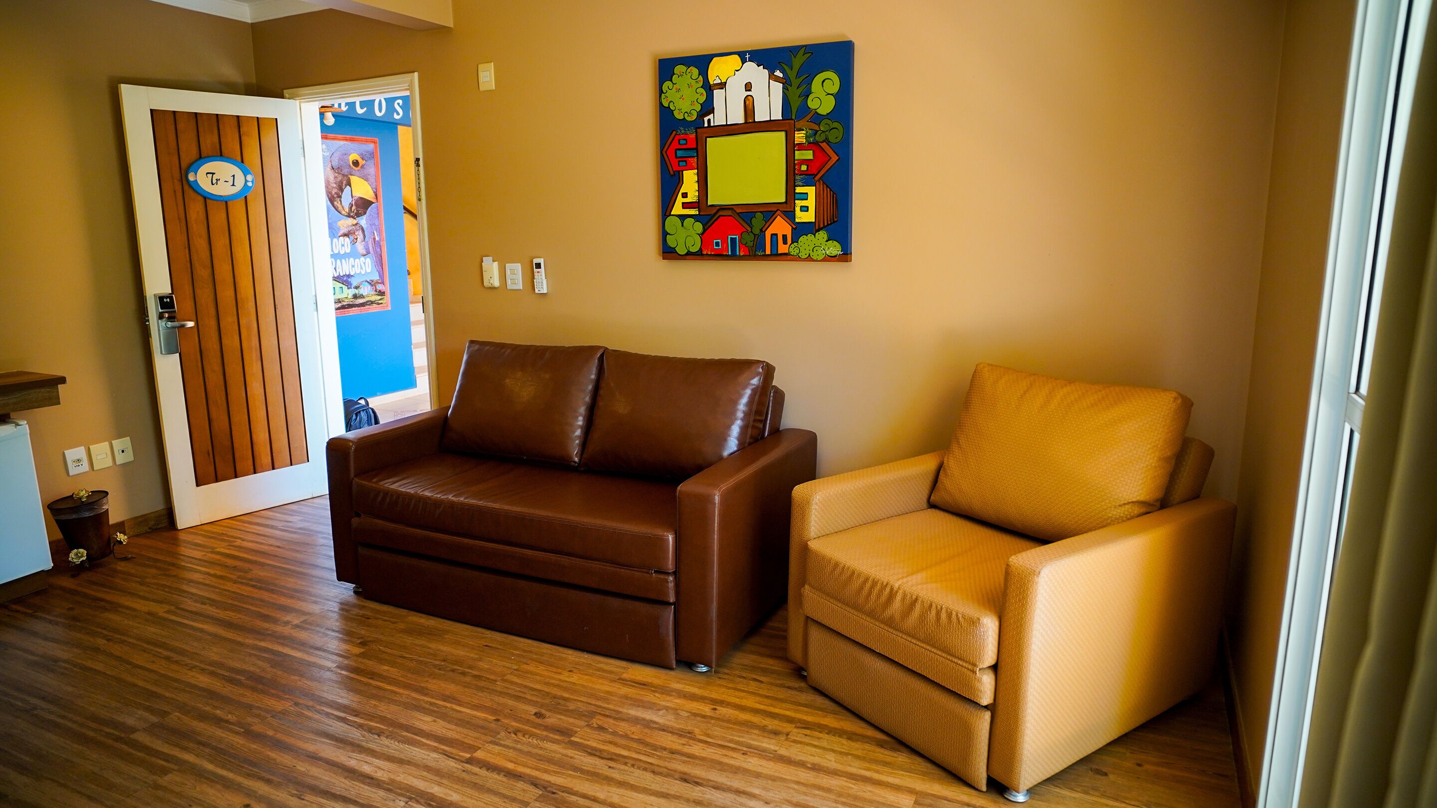 A room with a brown leather sofa, a tan armchair, wooden flooring, a door with a sign, and a colorful painting on the wall.