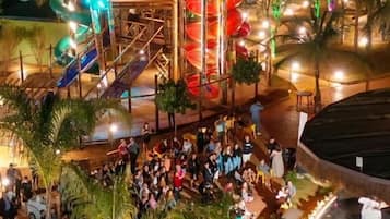 Ein Außenbereich bei Nacht mit einer zentralen Struktur, die eine Spiralrutsche und eine Sitzgelegenheit inmitten von Palmen beinhaltet.