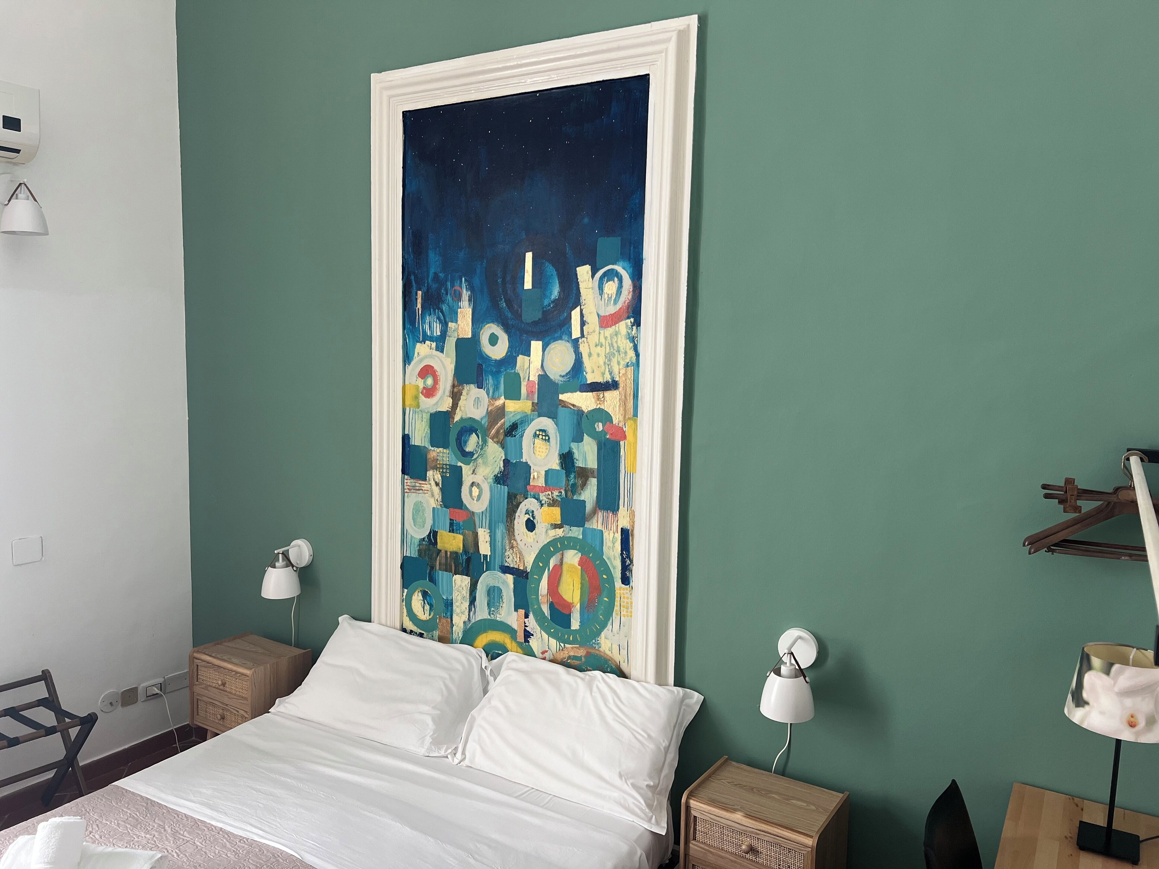 A bedroom with a bed, two bedside tables with lamps, a large abstract painting on the wall, and a wooden nightstand.
