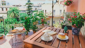 A wooden table with a plate of food, a book, and a drink on a balcony with potted plants and a view of buildings. - Bellini Home B&B (Catania)