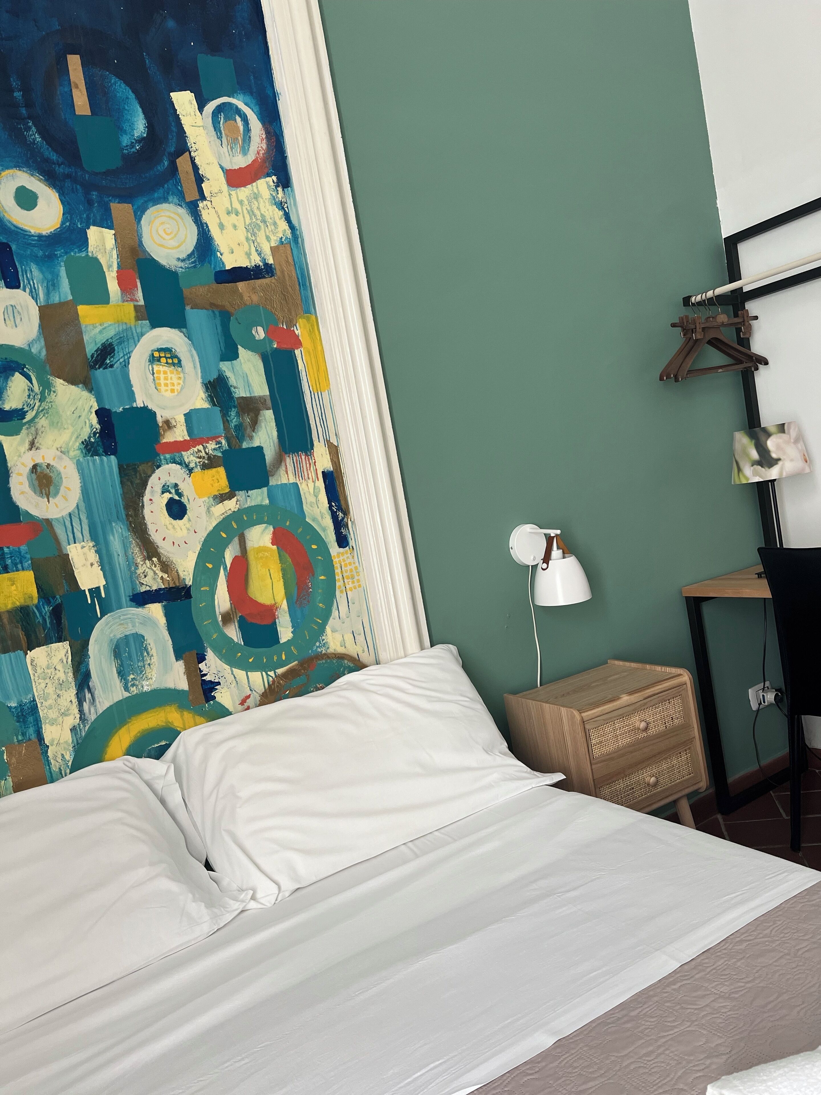A bedroom with a bed, a colorful abstract painting, a small wooden nightstand, a desk, and a wall-mounted lamp.