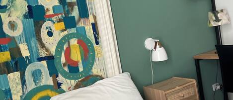 A bedroom with a bed, a colorful abstract painting, a small wooden nightstand, a desk, and a wall-mounted lamp.