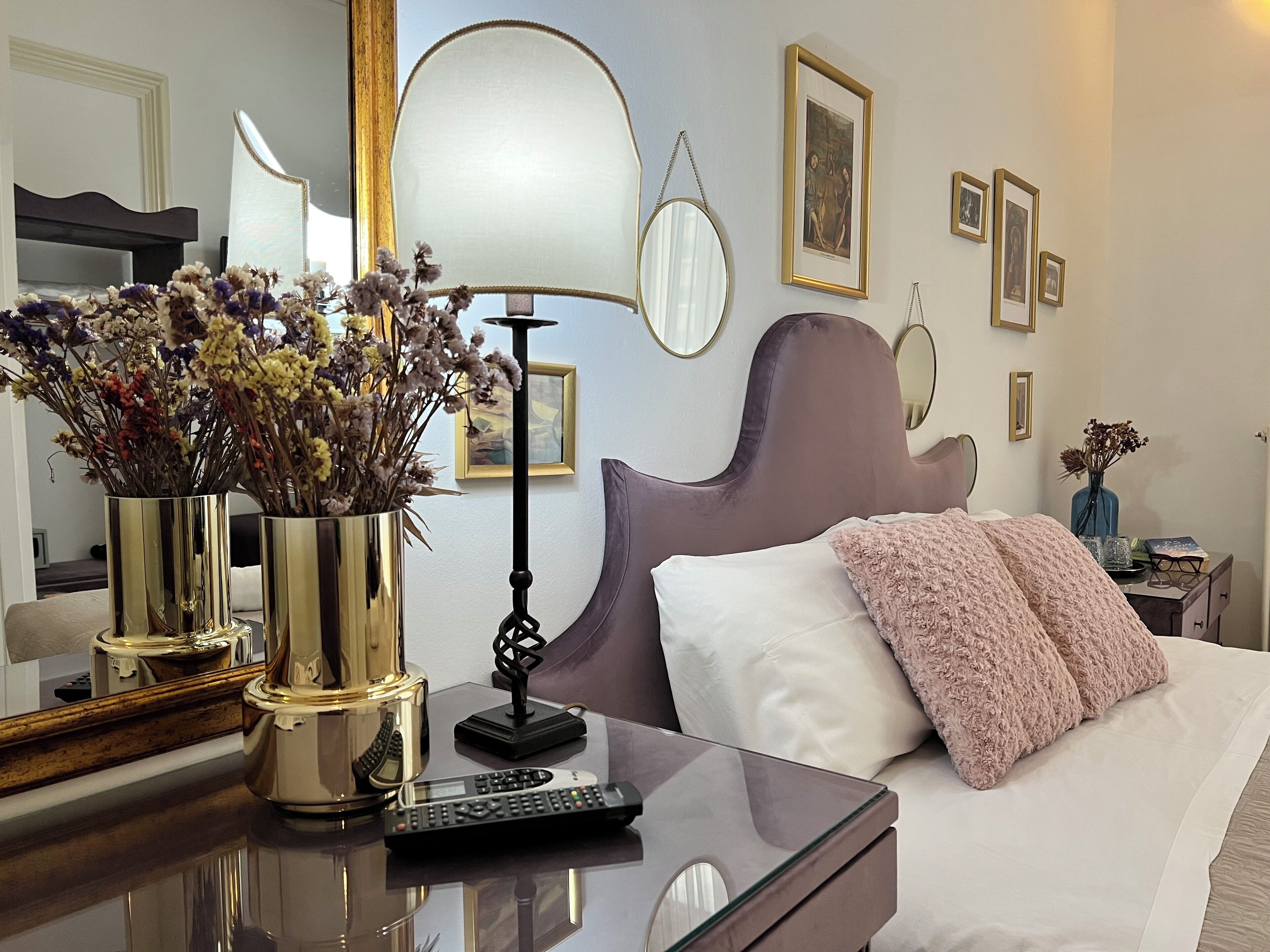 Ein Hotelzimmer mit einem Bett, einem Nachttisch mit Blumenvase, einer Lampe und einem Spiegel.