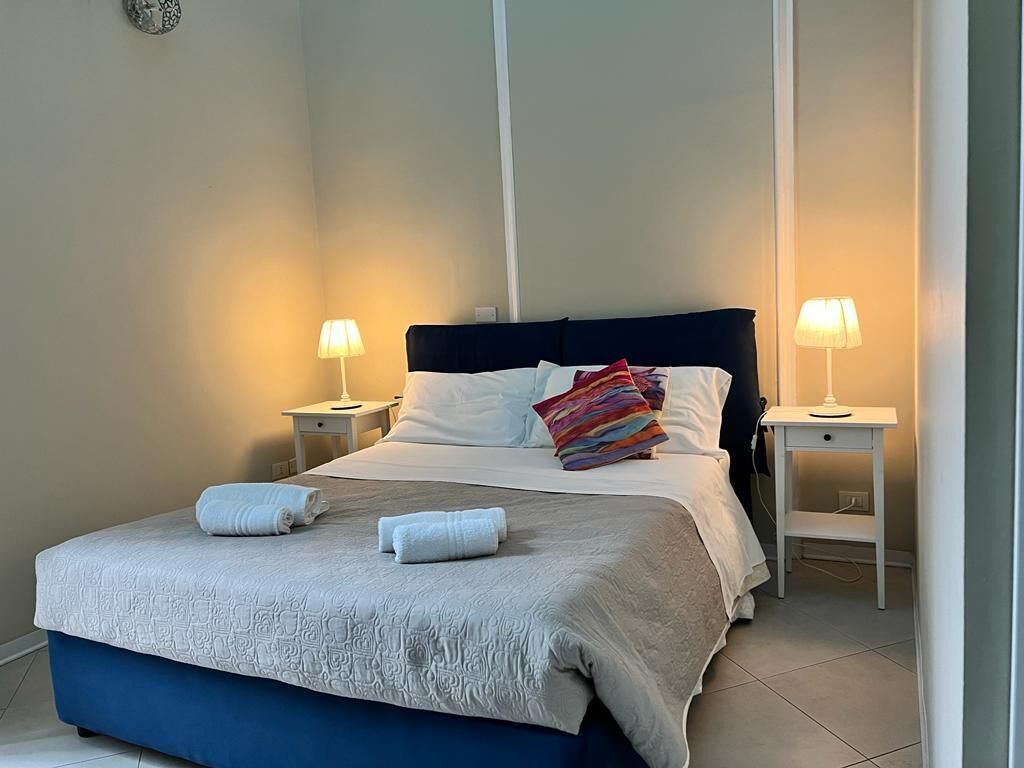 A neatly made bed with a white and blue bedspread, two white towels, and a colorful pillow. There are two bedside tables with lamps.