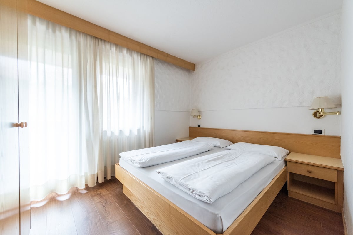 A bedroom with a wooden bed, a nightstand, a lamp, and a window with sheer curtains.