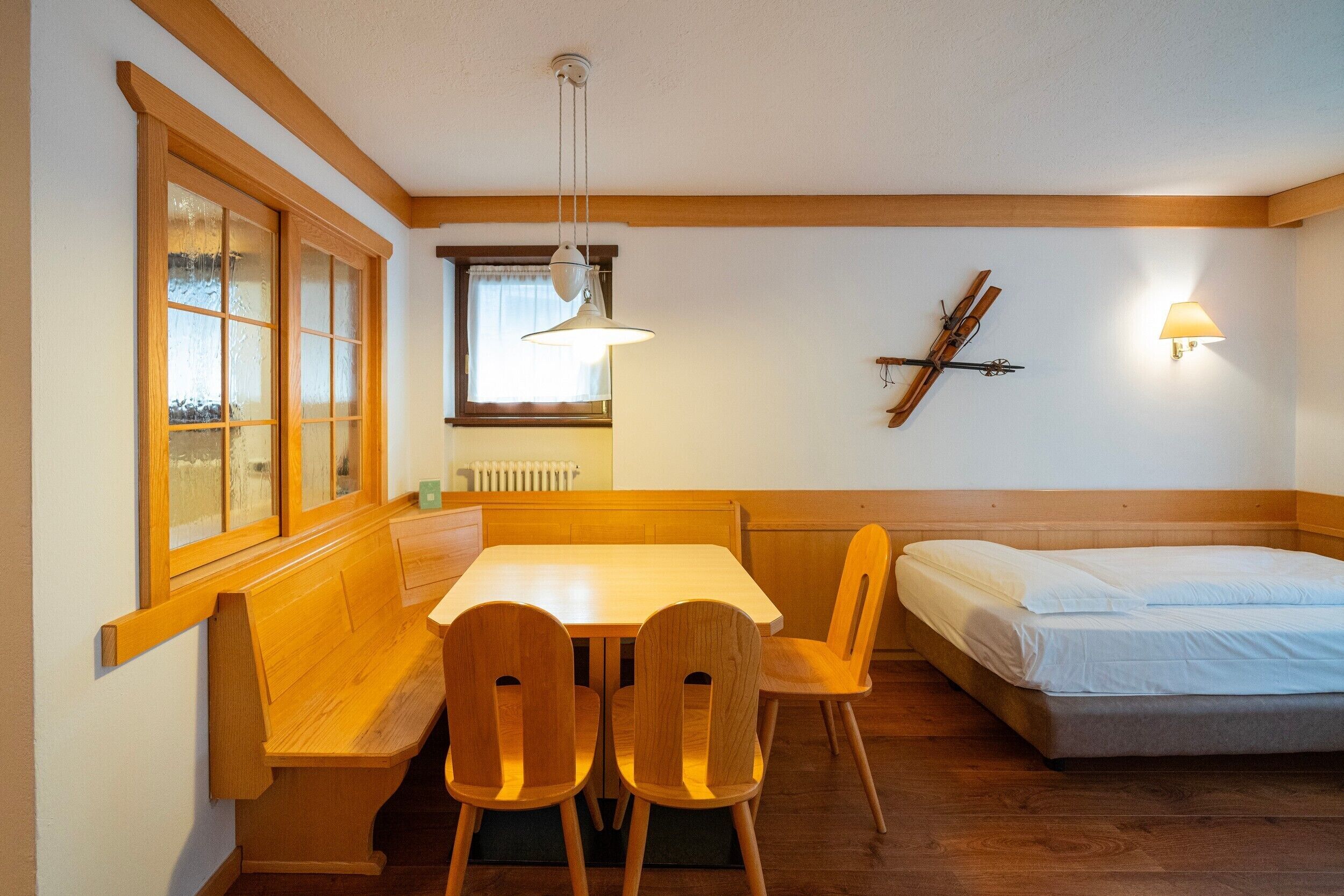 A room with a wooden bench, a table, and a bed. There is a window with frosted glass and a wall-mounted lamp.