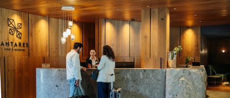 Un comptoir d’accueil avec un revêtement mural en bois et un plan de travail en pierre. Trois personnes se trouvent au comptoir et interagissent entre elles : l’une a une valise et l’autre, un sac.