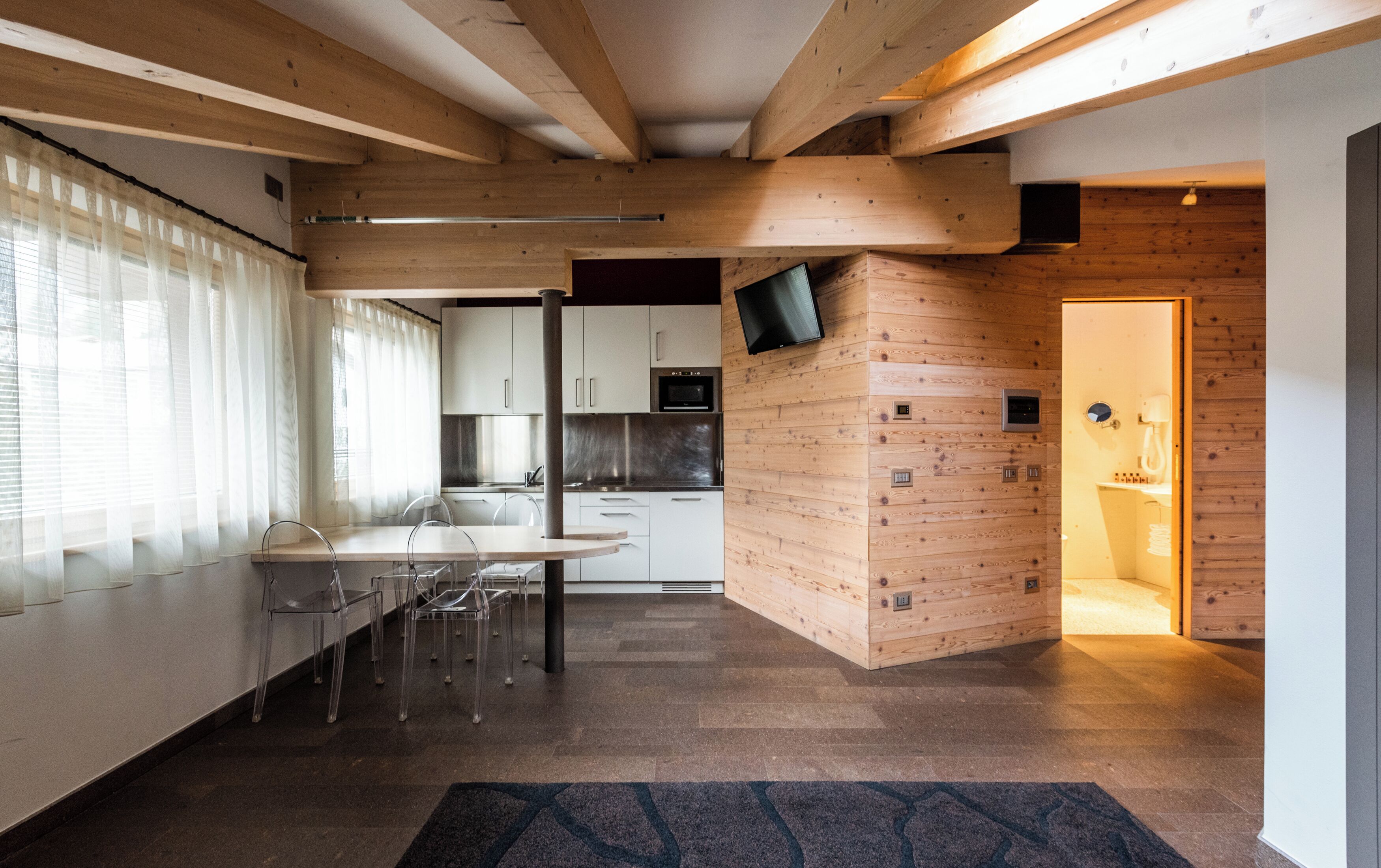 Una cucina moderna con travi in legno, una zona pranzo con sedie trasparenti e un bagno con lavandino a vista.