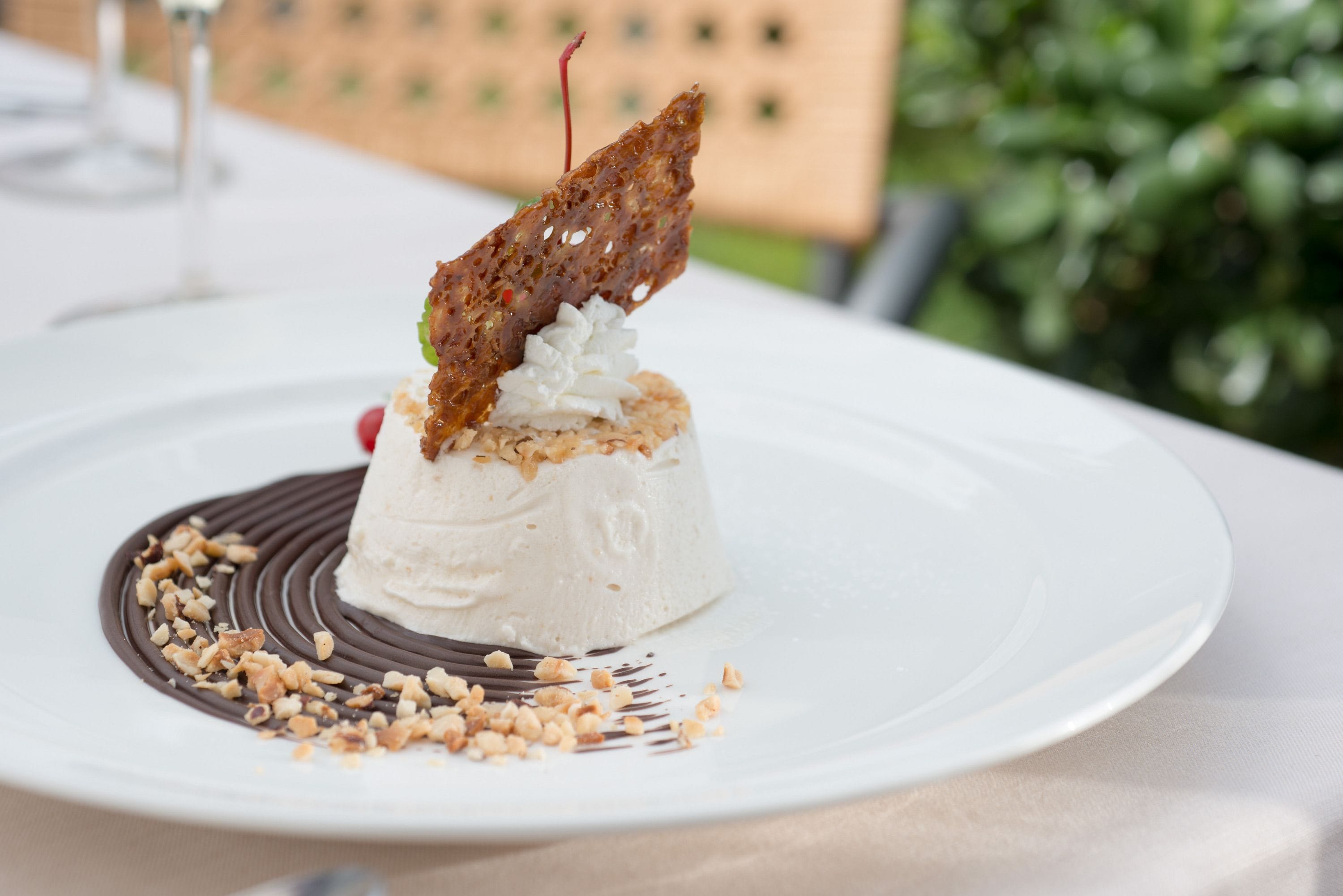 A dessert on a white plate with a chocolate design, nuts, and a piece of fruit on top.