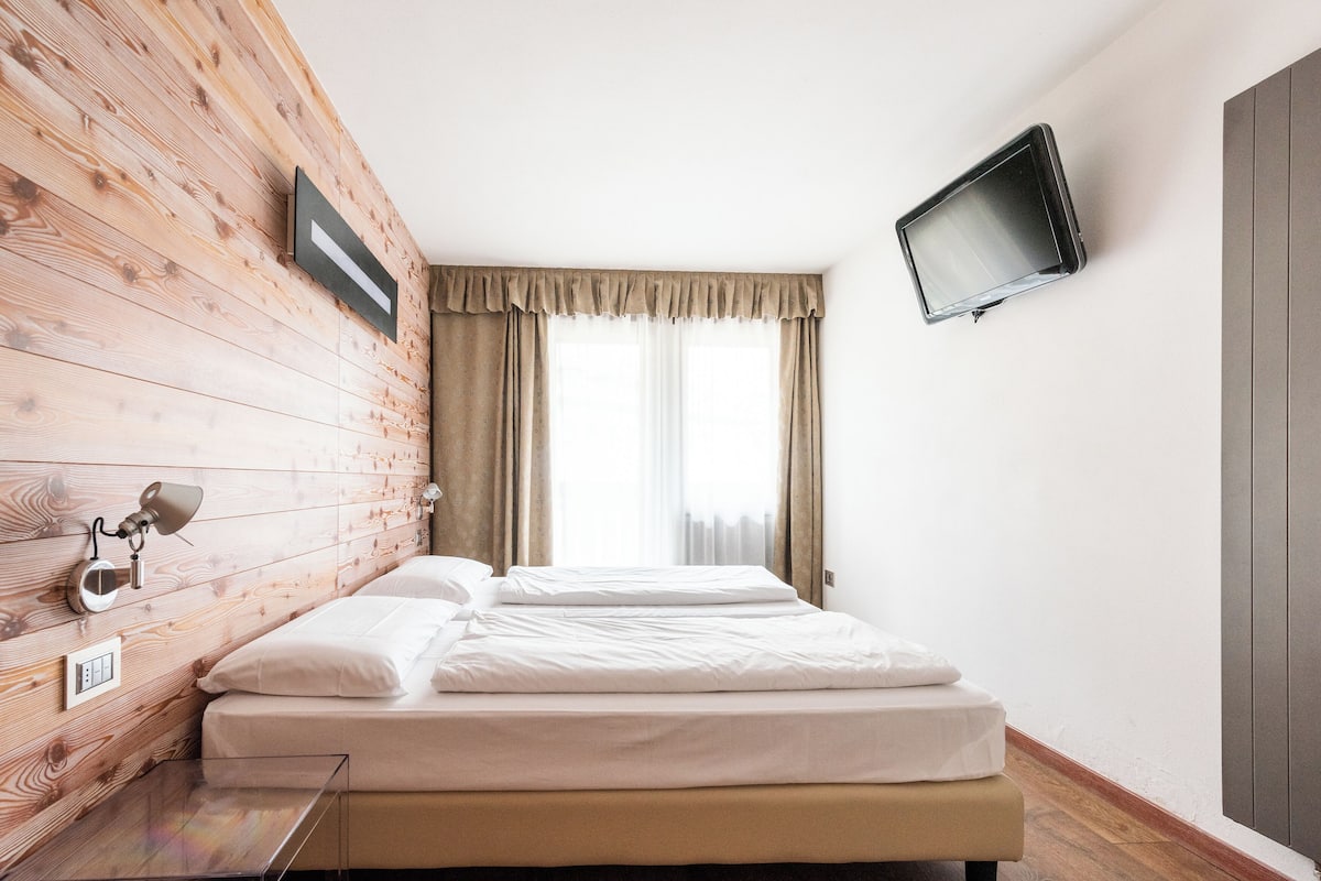 A hotel room with a wooden wall, a bed, a TV, and a window with curtains.