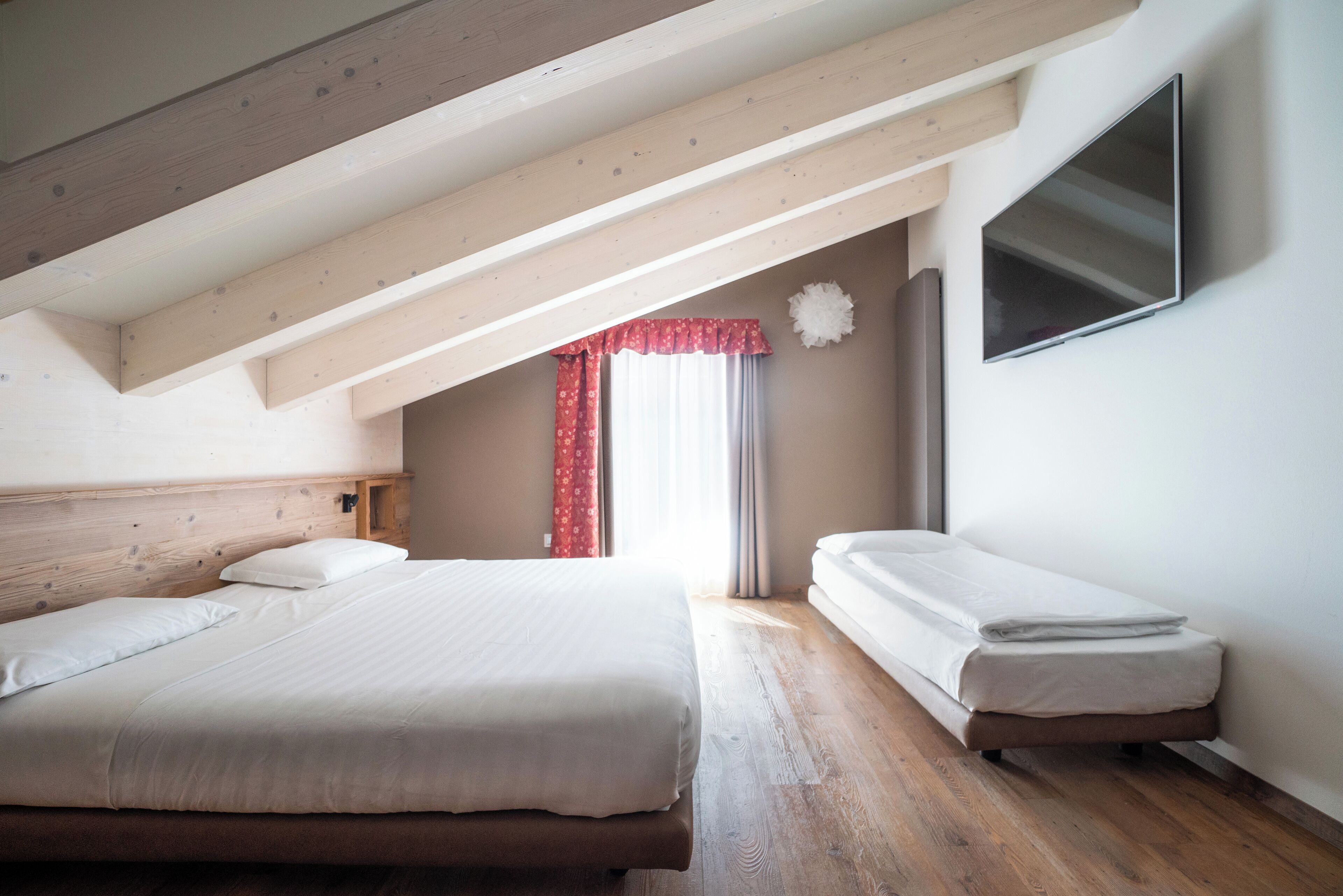 A bedroom with two beds, wooden beams, a TV, and a window with red curtains.
