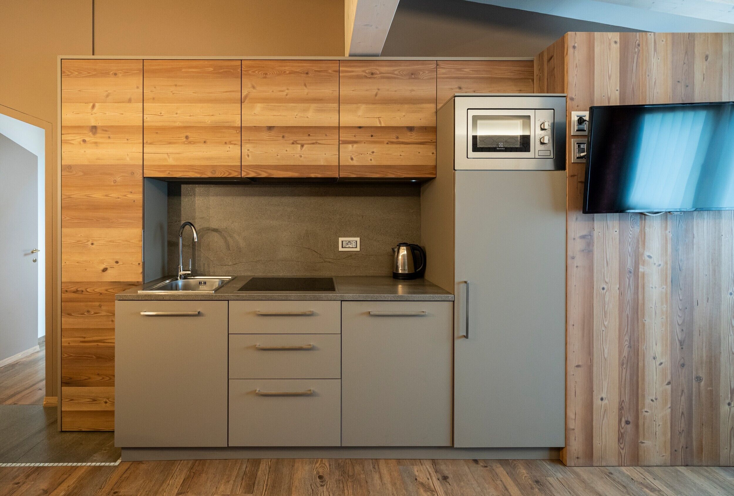 A compact kitchen with wooden cabinetry, a built-in oven, a coffee maker, and a flat-screen TV mounted on the wall.