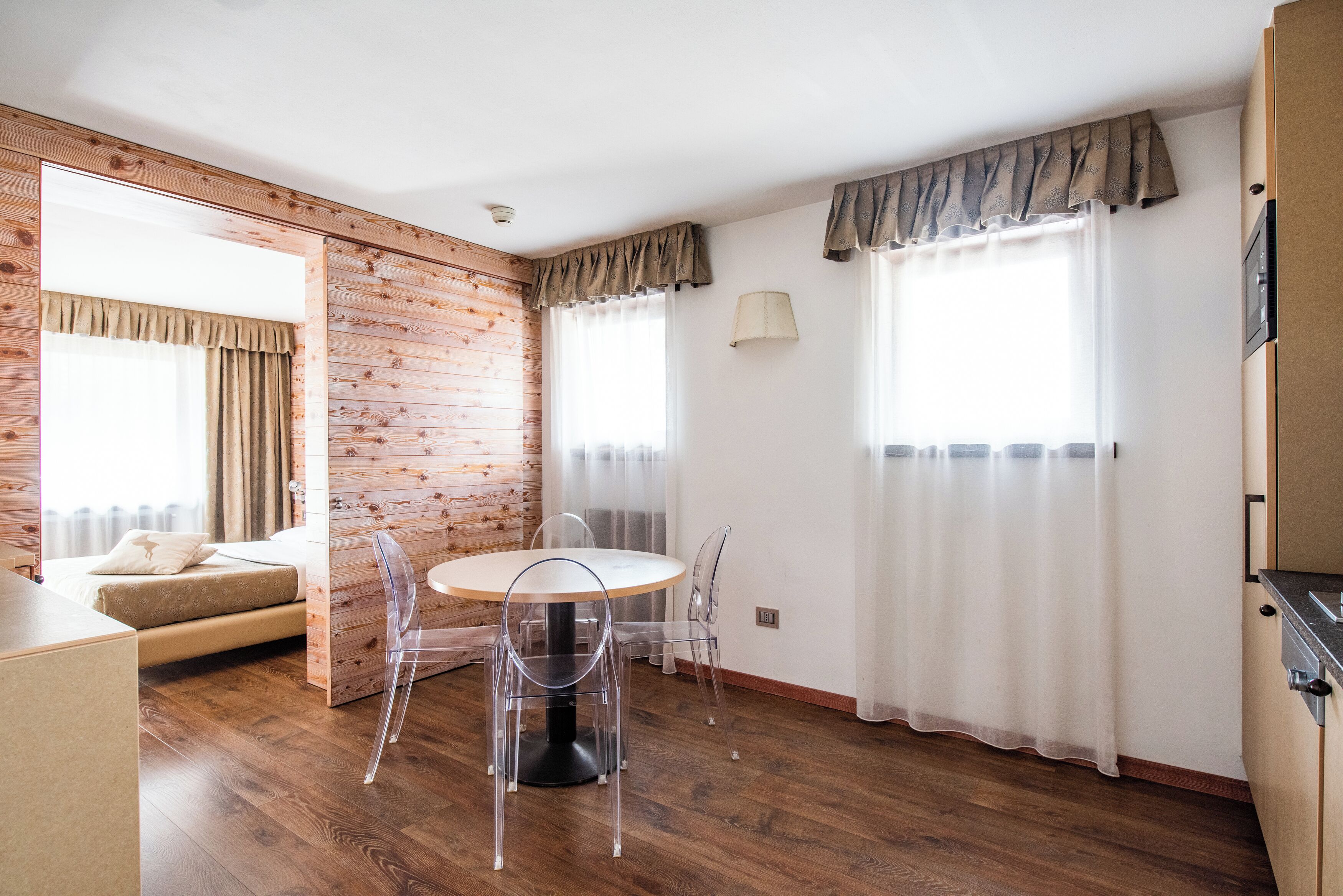 Une pièce avec un plancher en bois, une table ronde entourée de chaises, un lit recouvert d’une couette beige et une fenêtre avec des rideaux.