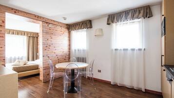 Une pièce avec un plancher en bois, une table ronde entourée de chaises, un lit recouvert d’une couette beige et une fenêtre avec des rideaux.