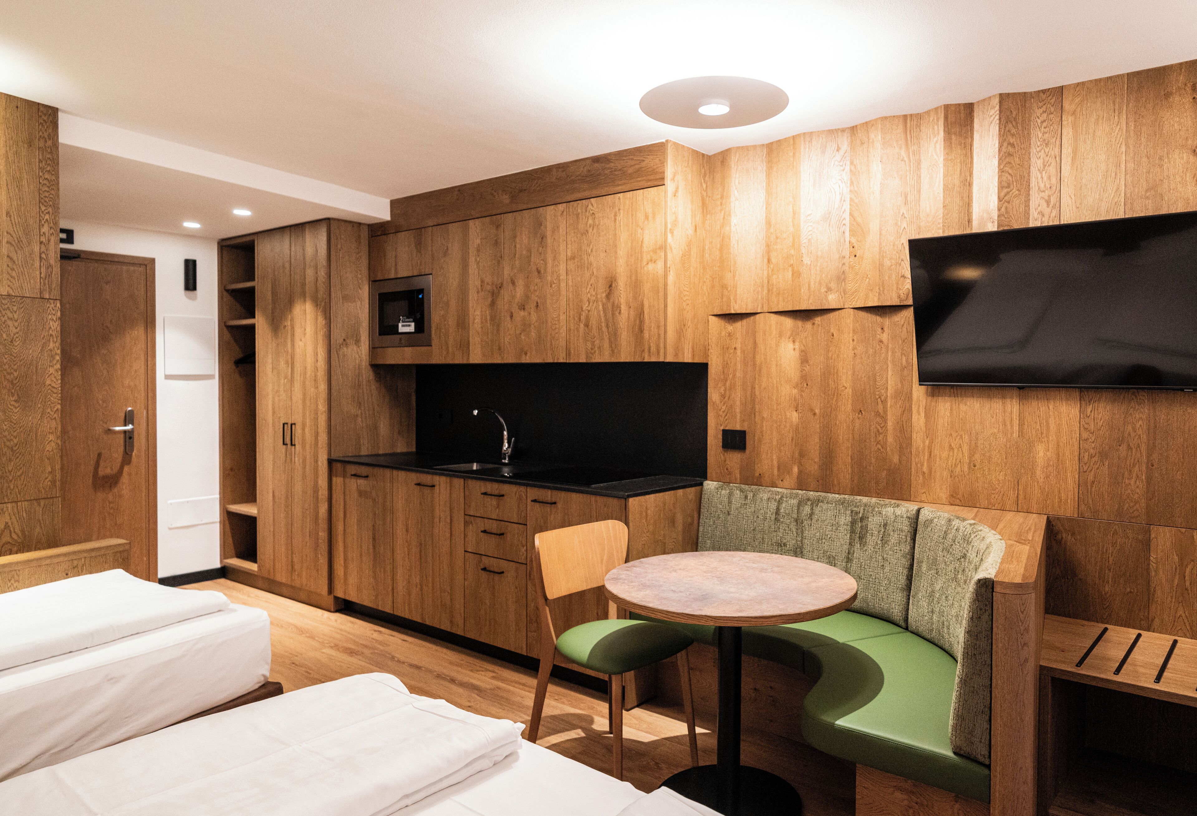 A compact room with a kitchenette, a flat-screen TV mounted on the wall, a round table with a green cushioned bench, and two single beds.