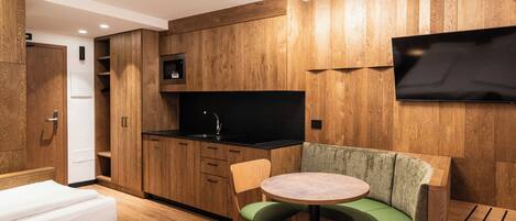 A compact room with a kitchenette, a flat-screen TV mounted on the wall, a round table with a green cushioned bench, and two single beds.