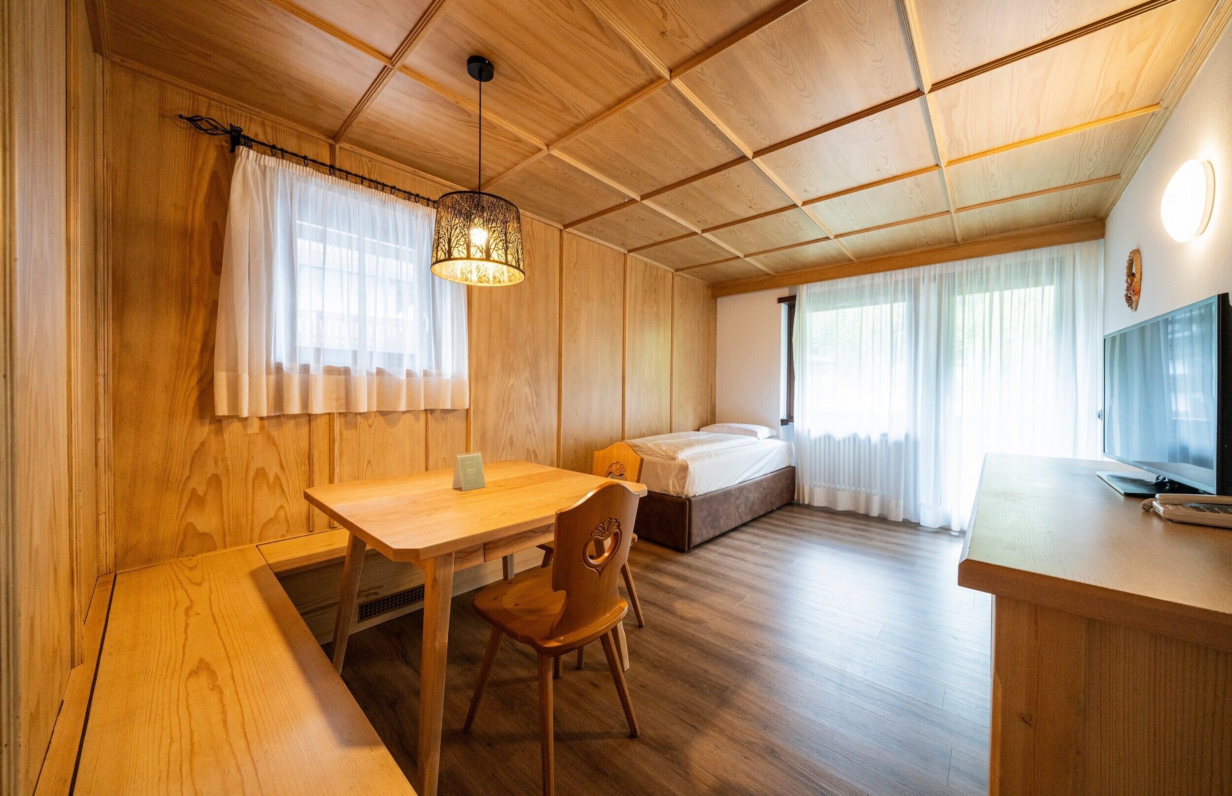 A room with a wooden ceiling, a bed, a desk, a chair, and a television.