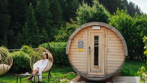Estructura de sauna de madera con puerta, rodeada de abundante vegetación y una terraza de madera.