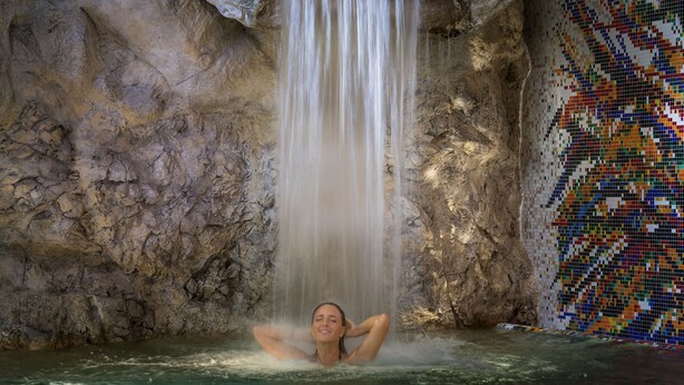 Una persona relajándose en una piscina con una cascada y un muro de mosaicos al fondo.