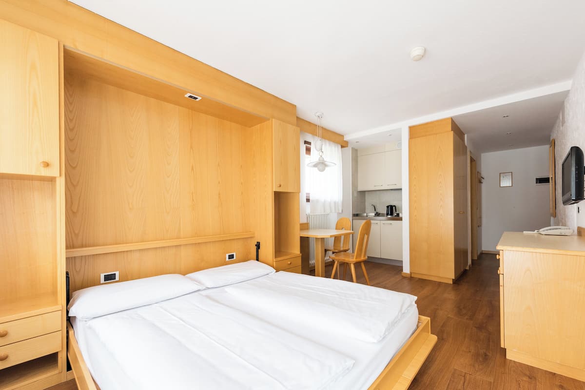A modern hotel room with a wooden bed, a small kitchen area, and a dining table.