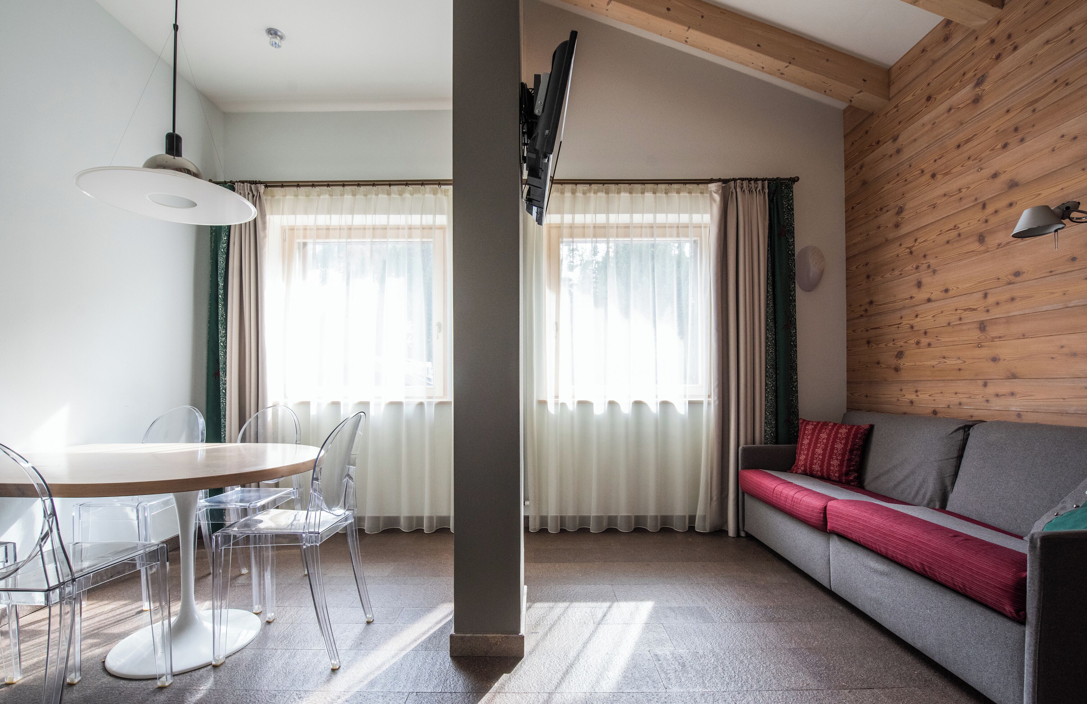 Een moderne woonkamer met een houten plafond, een ronde eettafel met transparante stoelen, een grijze bank met rode kussens en grote ramen met dunne gordijnen.