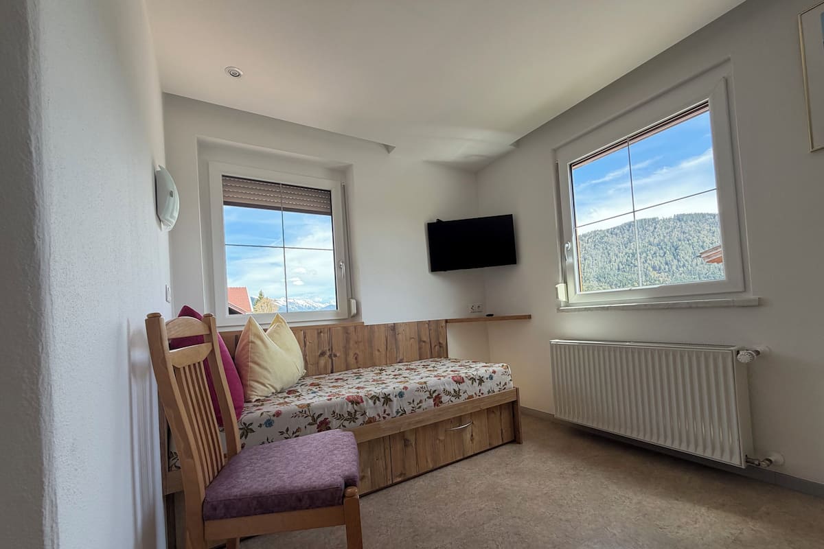 A small, cozy room with a bed, a chair, a TV, and a window with a mountain view.