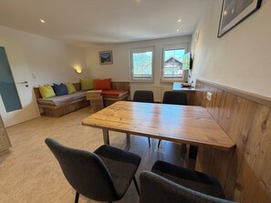 A room with a wooden dining table, two chairs, a wooden bench with cushions, a wall-mounted picture, and a window with a view of a house. - Haus Schönberg (Leutasch)